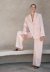 Vaaleanpunaisessa ylisuurassa puvussa oleva nainen seisoo yhdellä jalalla kaarevalla reunalla, mustat liukusandalit jalassa, yksinkertaista beigeä seinää vasten.