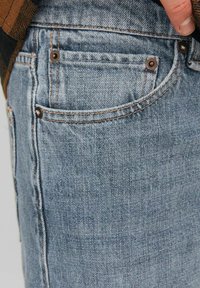 Jeans de denim azul claro con un corte clásico, costuras en contraste y remaches de cobre en los bolsillos. Textura suave con un acabado ligeramente desgastado.