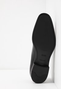 Chaussure habillée en cuir noir avec un bout pointu, une texture lisse et un accent minimaliste. Présente un logo embossé et une semelle en caoutchouc solide. Fabriquée en Italie.