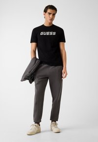 Zwart kort mouw t-shirt met "GUESS" logo, gecombineerd met grijze taps toelopende joggingbroek. Outfit bevat een grijs jack en lichtgekleurde sneakers.
