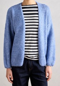 Gilet tricoté bleu clair, ouvert à l'avant, avec des manches longues et une texture douce, porté par-dessus une chemise à rayures noires et blanches. Jean foncé visible.
