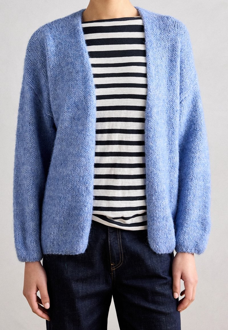 Gilet tricoté bleu clair, ouvert à l'avant, avec des manches longues et une texture douce, porté par-dessus une chemise à rayures noires et blanches. Jean foncé visible.