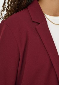 Blazer bordeaux en tissu texturé, avec un col à revers et une silhouette ajustée, superposé à un haut blanc côtelé et une chaîne en or.