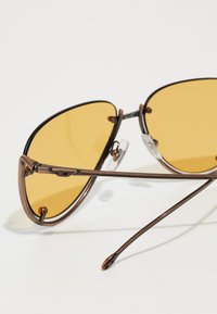 Occhiali da sole color bronzo con lenti gialle. Presentano una montatura in metallo elegante, appoggiatesti trasparenti e stanghette curve sottili.