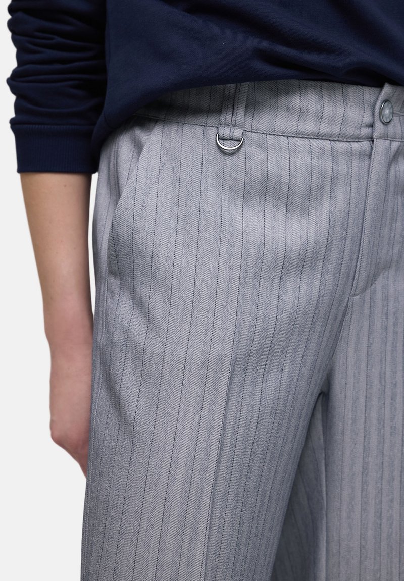 Personne portant un pantalon gris à fines rayures avec des poches latérales et un détail anneau en D sur la ceinture, associé à un haut bleu foncé.