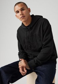Black hoodie made of cotton, featuring a raised "LEVI'S" logo on the chest. Worn with dark denim jeans. Positioned on a light wooden surface.