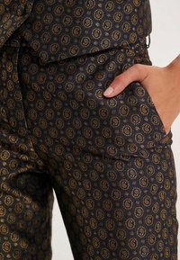 Pantalons à motifs sombres avec un design en tourbillon doré sur fond bleu marine, avec une poche latérale visible et une texture lisse.