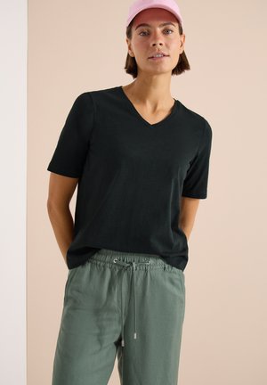 Femme portant un t-shirt noir à col en V, un pantalon vert à cordon et une casquette de baseball rose, debout avec les mains derrière le dos.