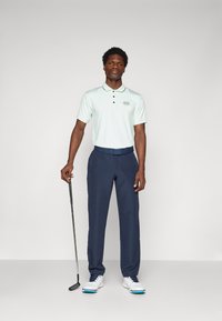 Ανοιχτό πράσινο polo shirt με μαύρα κουμπιά και λογότυπο, συνδυασμένο με ναυτικό παντελόνι και λευκά γκολφ παπούτσια με μπλε λεπτομέρειες. Κρατάει μια γκολφ μπαστούνι.