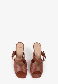 Brune syntetisk læder slingback hæle med krydsede stropper, firkantet tå og en struktureret mønsterbund. Har et guld spænde som accent.