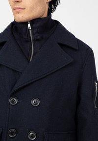 Granatowy wełniany płaszcz typu peacoat z podwójnym zapięciem na guziki, szerokimi klapami i kieszeniami bocznymi. Posiada ribbowaną wewnętrzną warstwę o wysokiej szyi z zamkiem błyskawicznym.