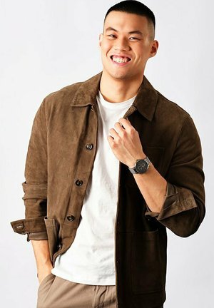 Braune Wildlederjacke mit Knopfverschluss, getragen über einem weißen T-Shirt. Das Modell zeigt eine silberne Uhr und präsentiert einen lässigen Look.