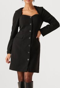 Zwarte jurk gemaakt van gestructureerde stof, met lange mouwen, een sweetheart halslijn, een knoopsluiting aan de voorkant en zijzakken.