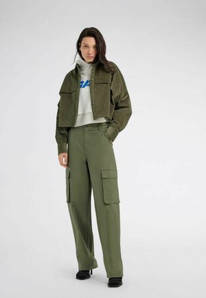 Donna che indossa pantaloni cargo verde oliva e giacca corta in velluto a coste sopra una felpa bianca con scritta blu, in piedi con una mano in tasca.