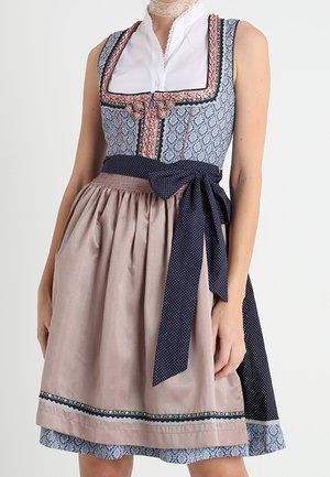 Femme portant un dirndl traditionnel avec un corsage bleu à motifs, une blouse blanche, un tablier rose pâle et une ceinture de taille marine à pois.