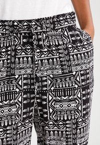 Pantalon noir et blanc à motifs avec une ceinture élastique, présentant des dessins géométriques et tribaux sur tout le tissu.