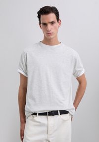 Young man wearing a light gray rolled-sleeve t-shirt tucked into white pants with a black belt, standing against a plain gray background.