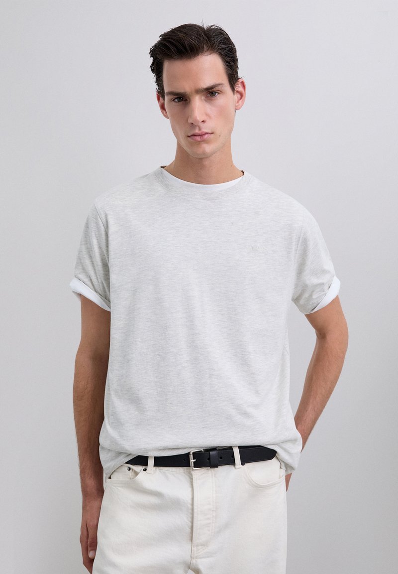 Young man wearing a light gray rolled-sleeve t-shirt tucked into white pants with a black belt, standing against a plain gray background.