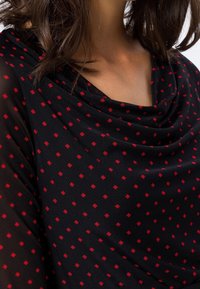 Femme portant un haut noir transparent à motif de petits carreaux rouges et un col drapé, montrant une partie du cou et de la poitrine.