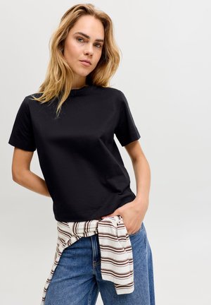 Γυναίκα με ξανθά μαλλιά που φοράει μαύρο μπλουζάκι και μπλε τζιν, με μια ριγέ πουκάμισο δεμένο γύρω από τη μέση της σε απλό φόντο.