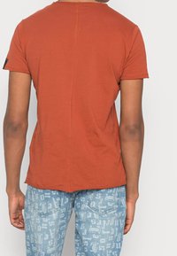Camiseta de manga corta color naranja oxidado con corte recto, que presenta una costura trasera y textura suave, combinada con vaqueros azul claro con estampado.