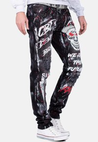 Jeans in denim nero con stampe grafiche, dettagli strappati, accenti a spruzzi rossi e bianchi, design a testa di teschio e decorazioni in borchie argentate.