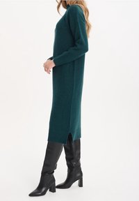 Tealfärgad stickad klänning med ribbade muddar, långa ärmar och sidoklyftor. Bärs med åtsittande svarta knähöga stövlar med spetsiga tår.