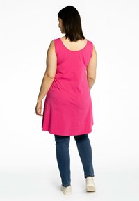 Roze mouwloze tuniek met een ronde hals, uitlopende onderrand en een ontspannen pasvorm, gecombineerd met donkerblauwe jeans en lichtgekleurde schoenen.