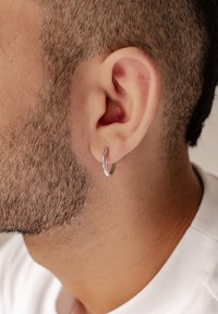 Silberner Creolenohrring im durchstochenen Ohr, mit einer glatten, reflektierenden Oberfläche. Kurze Haare sind eng geschnitten und zeigen die Hautstruktur.