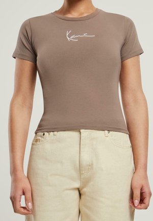 Bruine getailleerde t-shirt met korte mouwen en ronde halslijn, voorzien van witte cursieve tekst op de borst. Gecombineerd met lichtbeige jeans.