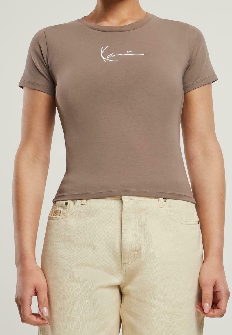 Brązowa dopasowana koszulka z krótkim rękawem i okrągłym dekoltem, z białym tekstem pisaną czcionką na piersi. Połączona z jasnymi beżowymi dżinsami.