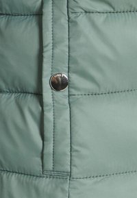 Sezione di giacca a vento verde chiaro, con bottone a pressione inciso con "ONLY", texture trapuntata e cuciture in contrasto.