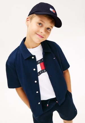 Gutt iført marineblå caps, kortermet skjorte med knapper og shorts over en hvit T-skjorte med logo, smilende med hendene i lommene mot hvit bakgrunn.