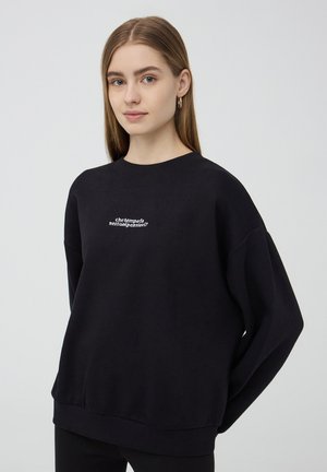 Felpa nera in morbido tessuto con scollo rotondo, spalle scese e testo ricamato bianco sul petto. Vestibilità rilassata.