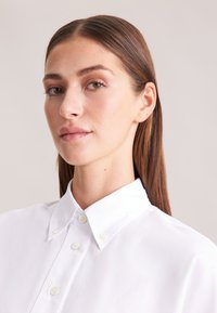 Camicia bianca con colletto classico, caratterizzata da una texture liscia e un leggero lucido, presentata su uno sfondo neutro.