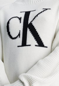 Maglione bianco a maglia a coste con un grande logo nero "CK" al centro. Le maniche sono larghe e presentano una texture a coste.