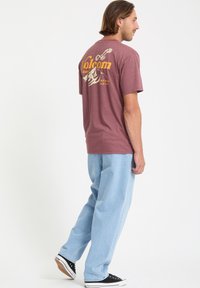 Bordeauxrotes T-Shirt mit Grafikdruck auf dem Rücken, featuring "Volcom"-Text und Bergdesign. Kombiniert mit hellblauen, lockeren Jeans und schwarzen Sneakers.