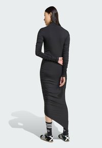 Svart långärmad klänning med samlad struktur, figurnära silhuett, hög krage och asymmetrisk fåll, tillsammans med svartvita randiga strumpor och skor.