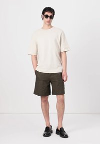 Beige košile s krátkým rukávem, olivově zelené kraťasy a černé slip-on boty. Model má černé sluneční brýle a krátké, strukturované vlasy.