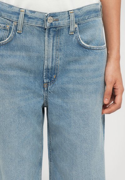 Jean en denim bleu clair avec taille haute, design à cinq poches, quincaillerie argentée et décoloration subtile. Le tissu a une finition lisse et texturée.