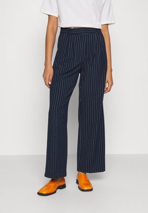 Persona que lleva pantalones anchos con rayas azul marino, una camiseta blanca corta y mocasines naranja brillante, de pie sobre un fondo liso.