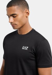 T-shirt in cotone nero con collo tondo e maniche corte, caratterizzato da un logo EA7 bianco sul lato sinistro del petto. Tessuto morbido, vestibilità casual.