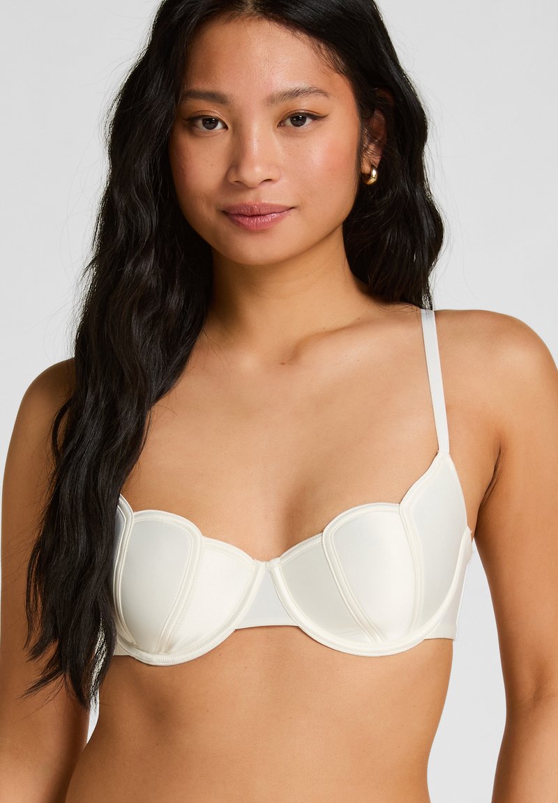 Kobieta z długimi czarnymi włosami, ubrana w biały biustonosz z fiszbinami i regulowanymi ramiączkami oraz złote kolczyki koła, zwrócona przodem na jednolitym tle.