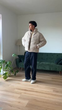 Giacca in pile beige imbottita con colletto alto. Indossata sopra un top beige chiaro, abbinata a pantaloni blu scuro e sneakers bianche. Ambientazione interna.