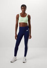 Mintgroene sportbeha, marineblauwe legging met witte zijstrepen, witte sport schoenen. Materiaal lijkt rekbaar; aansluitend ontwerp voor beweging.