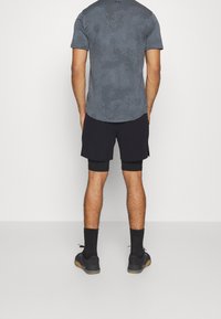 Maglietta atletica grigia con un motivo a pois e maniche corte, abbinata a pantaloni corti neri e sneakers scure, in piedi su uno sfondo semplice.