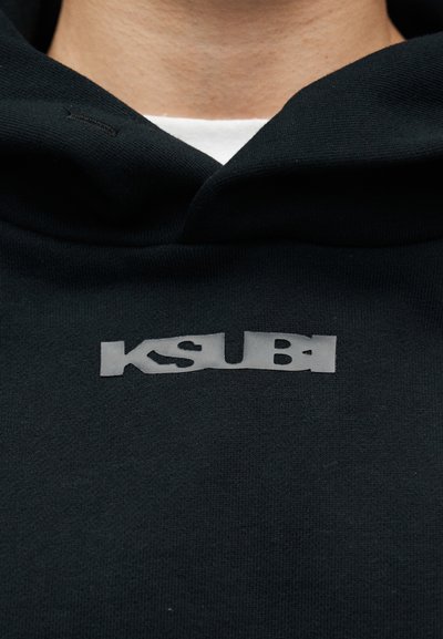 Μαύρο φούτερ με κουκούλα και ανάγλυφο, αντανακλαστικό λογότυπο KSUBI τυπωμένο στο στήθος. Κατασκευασμένο από απαλό, ελαστικό υλικό.