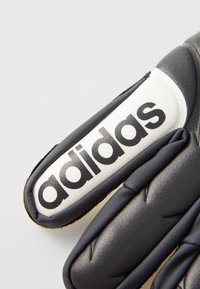 Gant en synthétique noir avec un design rembourré, présentant une étiquette blanche affichant le logo "adidas" en lettres noires épaisses.