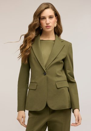 Kobieta z długimi, falowanymi włosami ubrana w oliwkowo-zielony, dopasowany żakiet i pasujące spodnie, stojąca na tle jasnego, jednolitego tła.