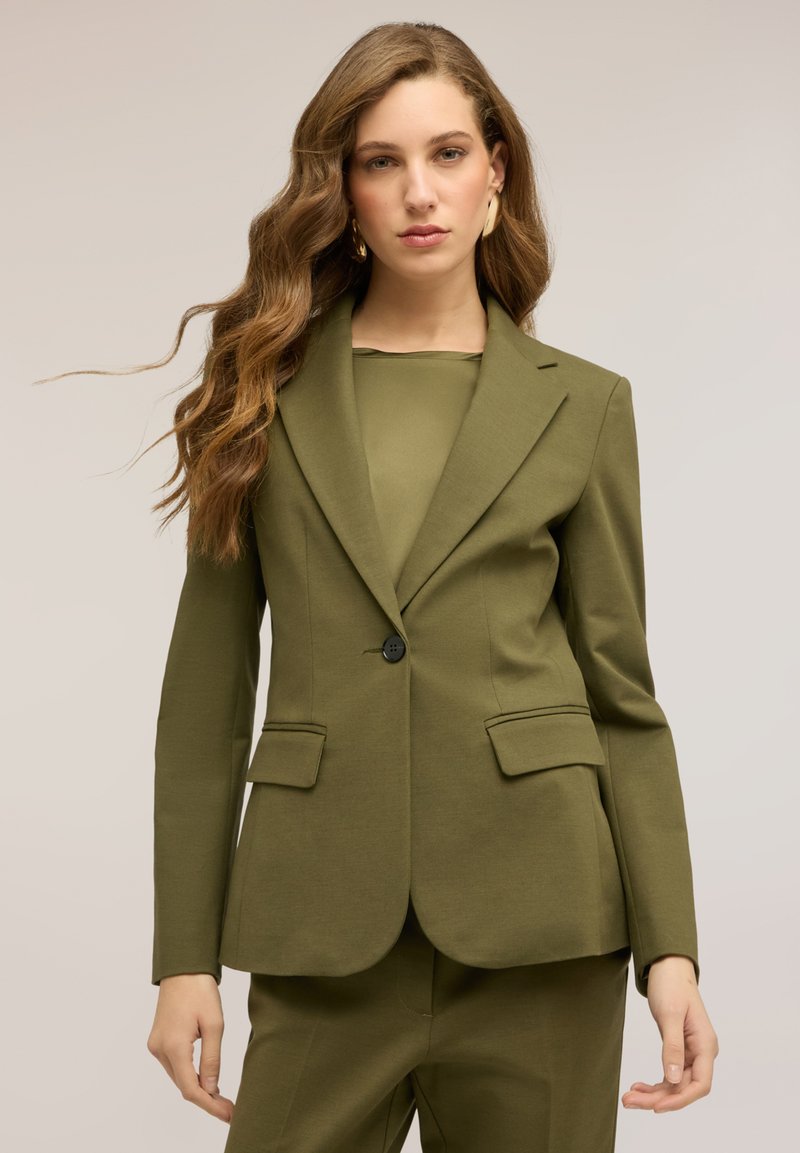 Vrouw met lang golvend haar, gekleed in een olijfgroen op maat gemaakt colbert en bijpassende broek, staande tegen een effen lichte achtergrond.